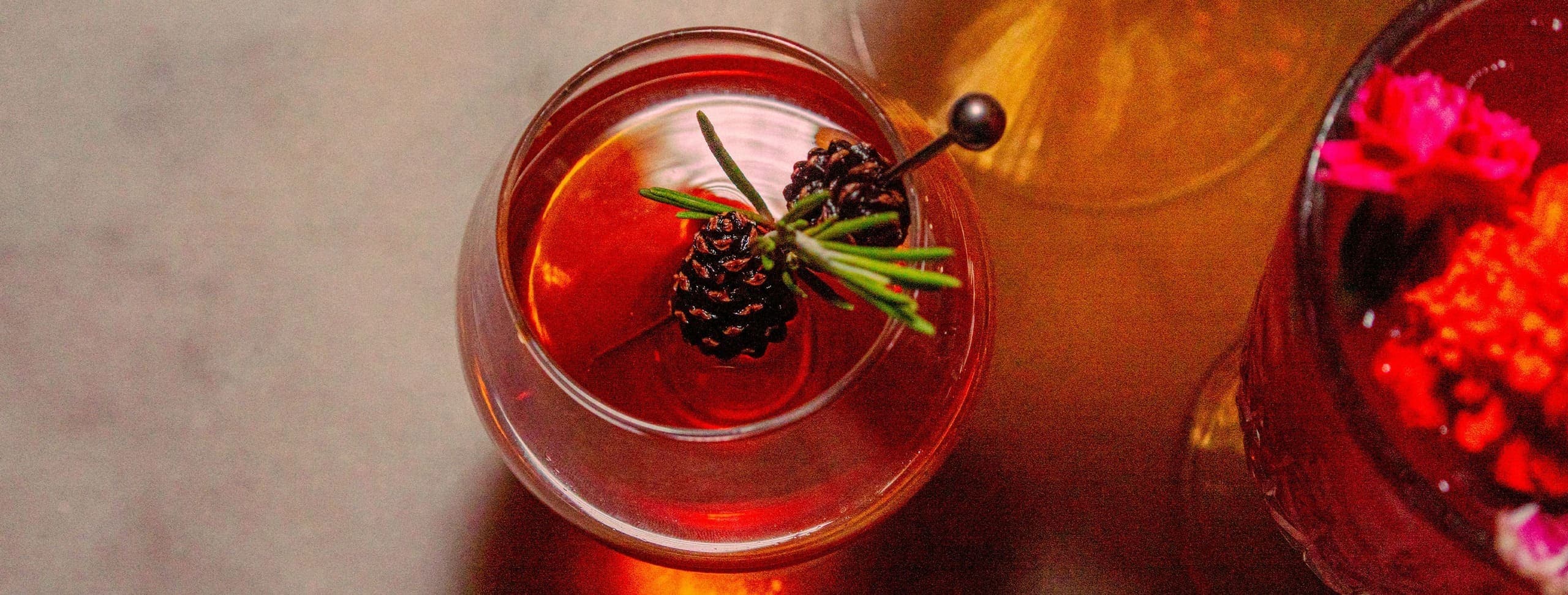 Red cocktail garnished with pine cone and herb sprigs on marble surface
